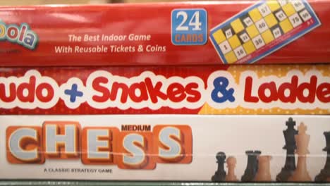 A-close-up-pedestal-shot-of-the-boxes-of-board-games-stacked-on-a-shelf-in-a-toys-store,-games-include-crossword,-Chess,-Snakes-and-ladders