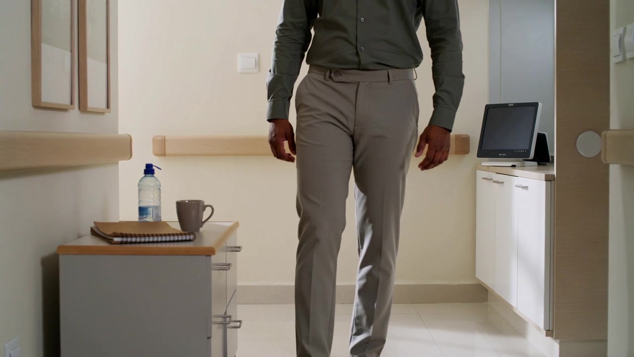 Entrando na clínica, homem caminhando vestindo camisa de botão, movendo-se em direção à câmera para usar tablet, com notebook.