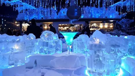 colorful cocktails rest on an ice bar counter as a bartender prepares drinks at an outdoor winter venue, surrounded by snow and illuminated by festive lights during a lively night