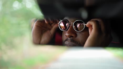 Car-Rear-Mirror-Reflection-of-African-Man-Putting-on-Sunglasses-Sitting-Inside,-Face-close-up