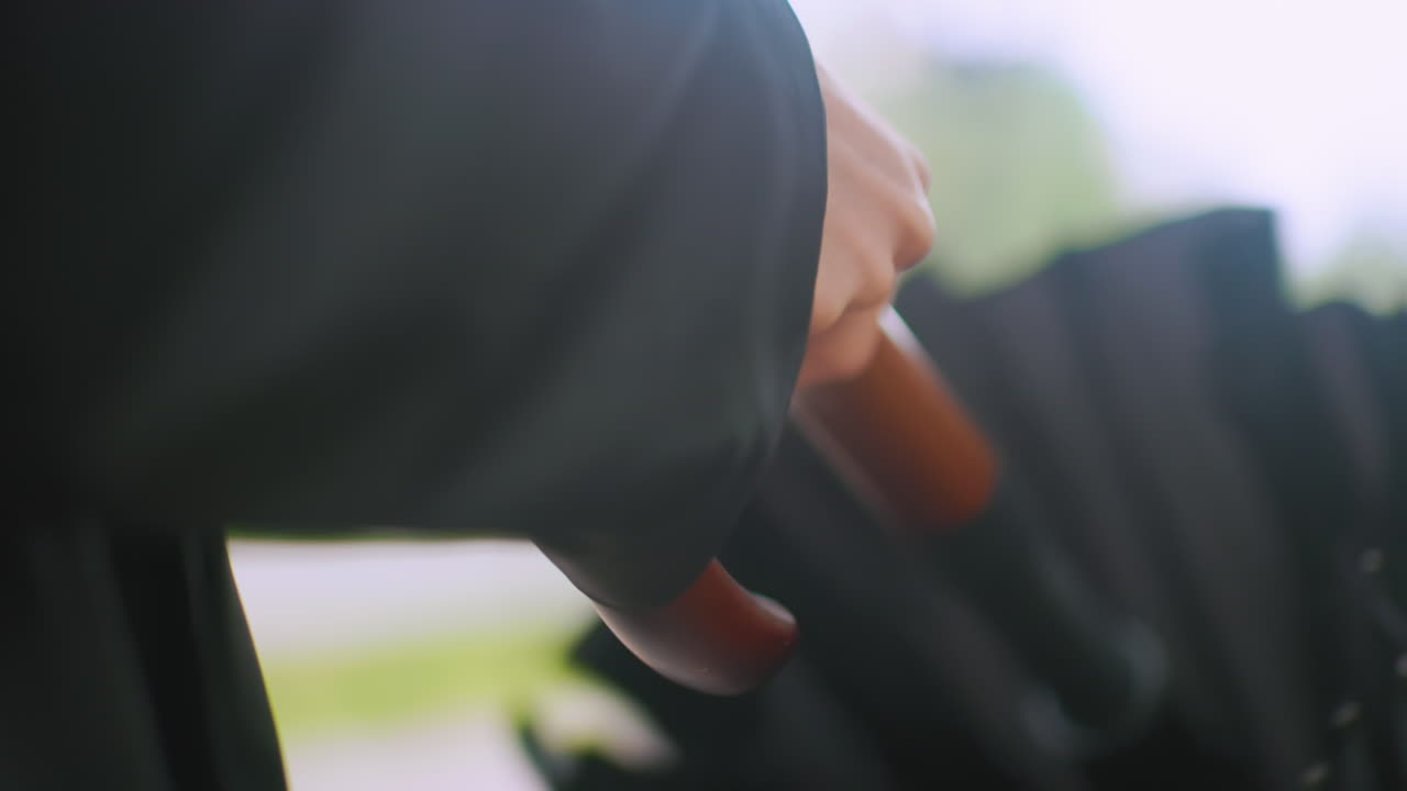 Close up of hand holding wooden handle of closed black umbrella outdoors on rainy day with blurred green background symbolizing protection, waiting, anticipation, weather, preparation, resilience