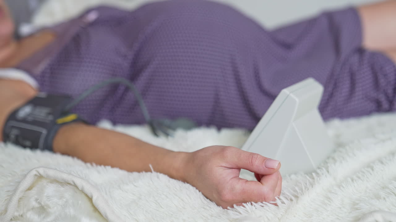 Pregnant woman measuring her blood pressure while calmly lying on the bed. Pregnant lady on the bed in blur. Black cat walking in the background.
