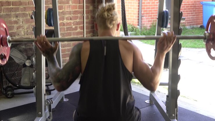 Muscly man in home gym exercising barbell squat shot from back