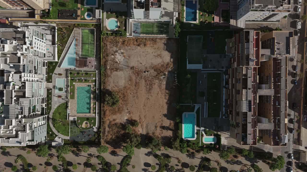 Birds eye view of an empty plot of land surrounded by buildings with swimming pools, suggesting urban development and planning