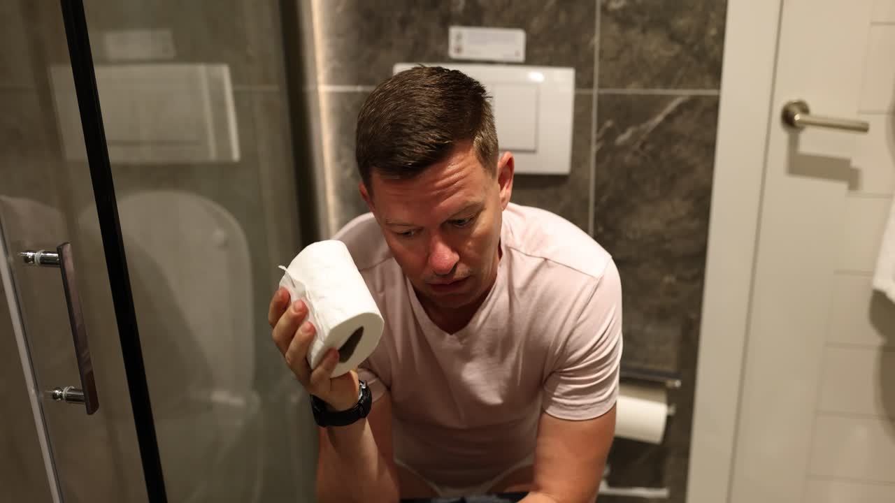 A man in a bathroom looking sad while holding a roll of toilet paper