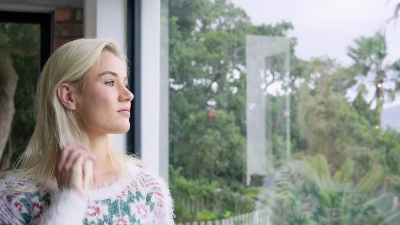 Woman brushing hair and gazing at fence through floor-to-ceiling window in living room, copy space