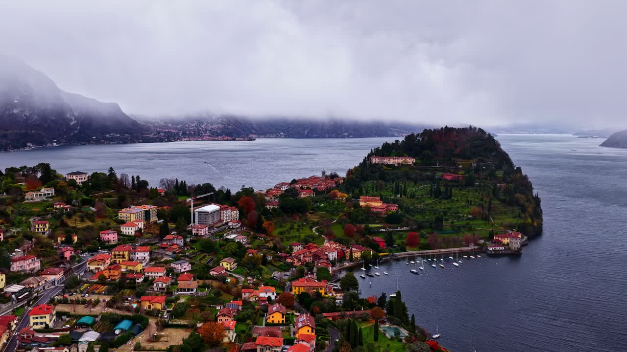 Vista aerea nebbiosa di Bellagio sul Lago di Como, che mostra le colorate case sulla collina, la tenuta con giardino nella penisola, l'acqua calma e le nuvole basse che si spostano sulle montagne circostanti