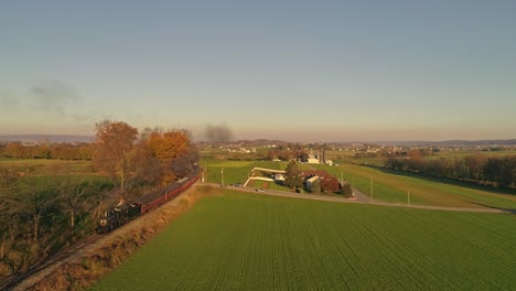 vista aérea de una antigua locomotora de vapor que se acerca tirando de los coches de pasajeros y soplando humo y vapor durante la hora dorada a última hora de la tarde