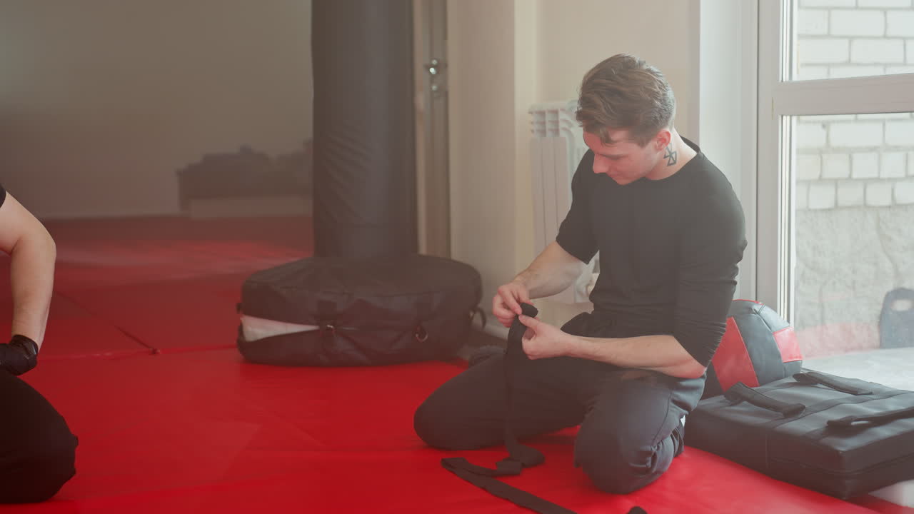 Athlete kneels on red mat near window preparing training hand wraps, focused and concentrated before workout session, dressed in black sportswear, showing dedication, strength, and discipline in martial arts
