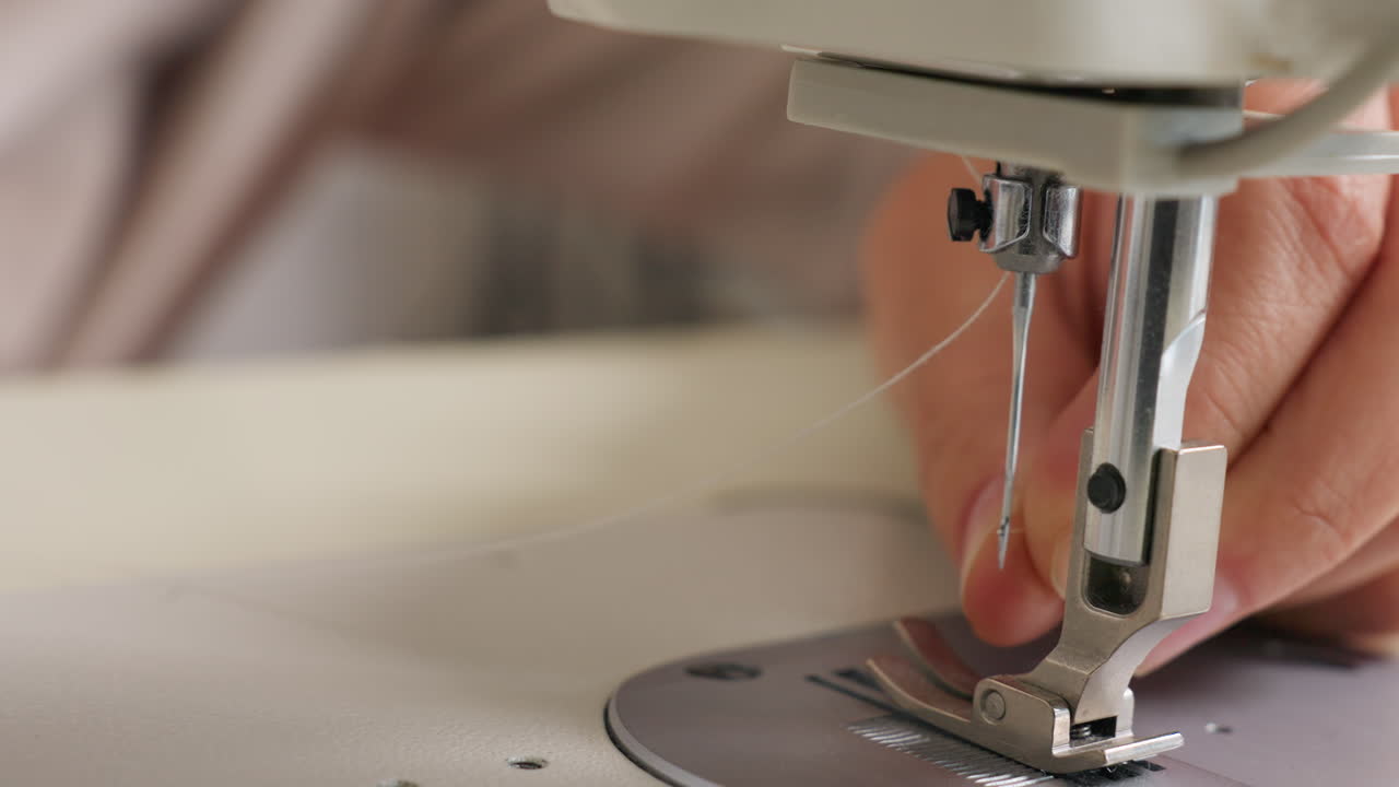Garment maker hand carefully passing sewing strand through needle, capturing skill, detail, gentle tension in minimal workshop environment, highlighting technique for precise finishing