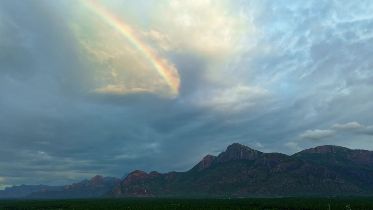 Pelangi melengkung di antara awan badai di atas pegunungan terjal di Tamil Nadu, India Selatan, perkebunan kelapa lebat di bawahnya, cahaya monsun dramatis, rekaman drone sinematik pemandangan India yang indah