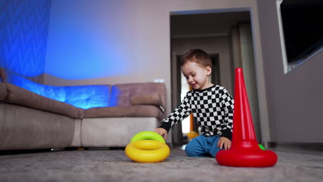 lovely caucasian toddler sits on the floor near the red conus. cheerful baby puts a ring on the pyramid base. low angle view.