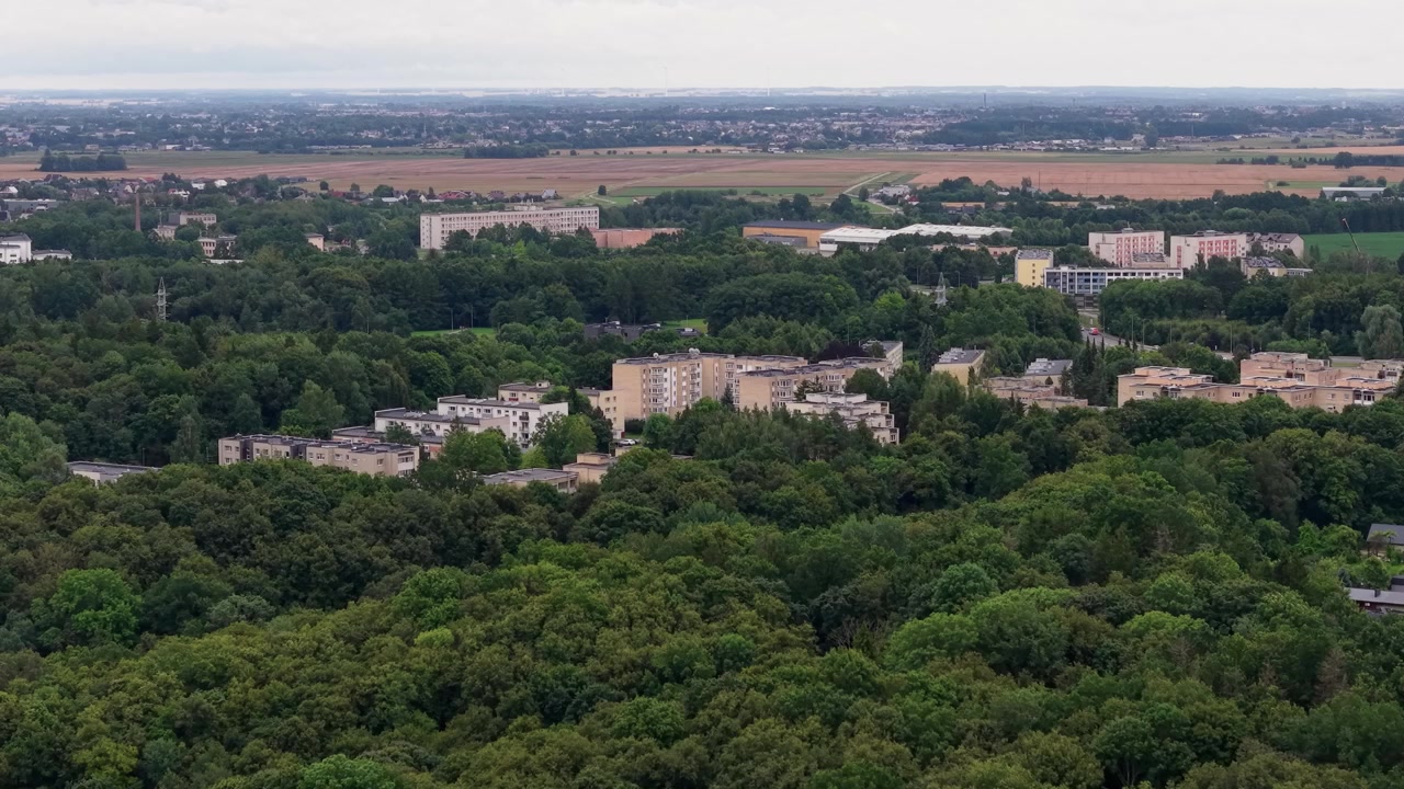Dronevideo vliegend over appartementencomplexen genesteld in weelderige groene bossen, met uitgestrekte landerijen daarachter. Een bewolkte hemel zorgt voor zacht, natuurlijk licht, ideaal voor schilderachtige landschapsbeelden.