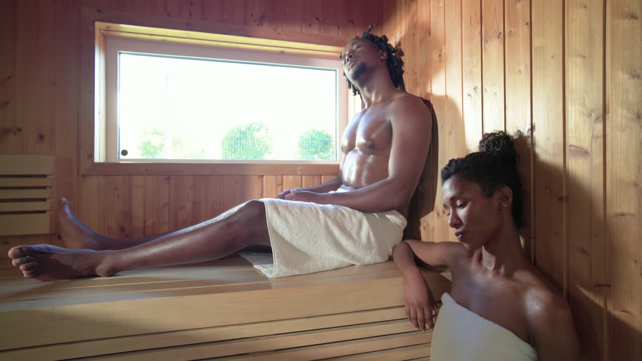Amici afroamericani che reagiscono alla luce dalla finestra, spostandosi e rilassandosi sulle panche della sauna avvolti negli asciugamani.