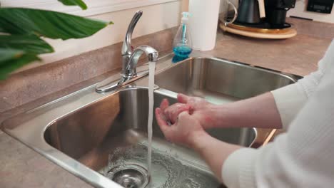 a young caucasian man washes his hands in a modern stainless steel natural light kitchen sink with a granit countertop blue soap and appliances