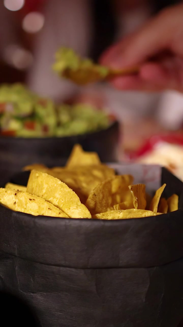Una mano afferra un nacho da un cestino e lo intinge nel guacamole in un ambiente di ristorante con illuminazione calda, con bassa profondità di campo.