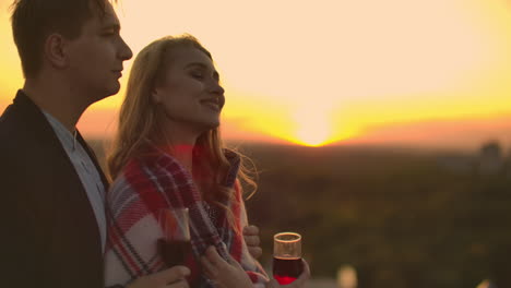 Liebende-Umarmen-Einen-Mann-Und-Ein-Mädchen-Und-Beobachten-Den-Sonnenuntergang-Bei-Einem-Glas-Wein,-Während-Sie-Auf-Dem-Dach-Eines-Gebäudes-Stehen.-Zeitlupenbild-Der-Beziehung-Eines-Jungen-Ehepaares.