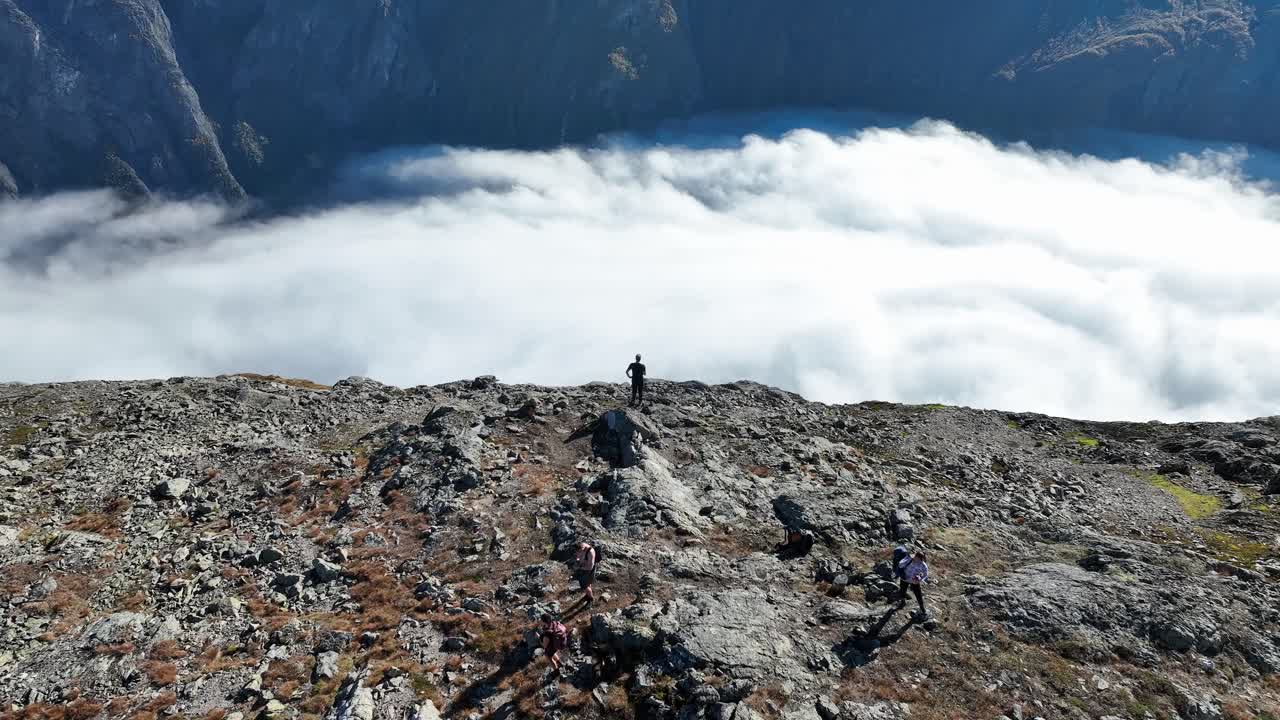 Slow aerial orbit of a man standing on a mountain peak above Naeroyfjord, with morning clouds over the fjord and other hikers visible below