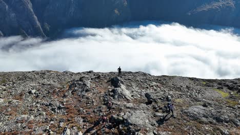 slow aerial orbit of a man standing on a mountain peak above naeroyfjord, with morning clouds over the fjord and other hikers visible below