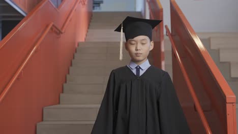 portrait of a happy asian preschool male student in cap and gown holding graduation diploma and looking at the camera