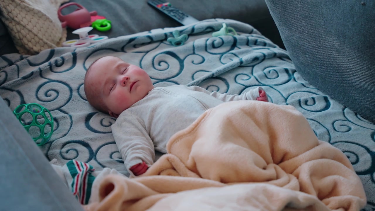 Inquadratura fissa da dietro un divano che mostra un neonato che dorme, coperto da una coperta e che indossa una tutina grigia all'interno di una casa di famiglia