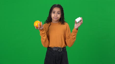 unsure african american teen girl on green screen holding drugs and orange shrugging shoulders. portrait of uncertain teenager choosing pharmaceutical medication or healthful fruit on chromakey.
