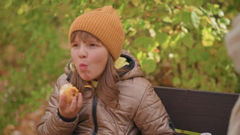 young girl in warm jacket and orange beanie taking joyful bite of apple while sitting on bench, smiling with eyes closed, adult figure beside her observing gently in serene autumn park setting