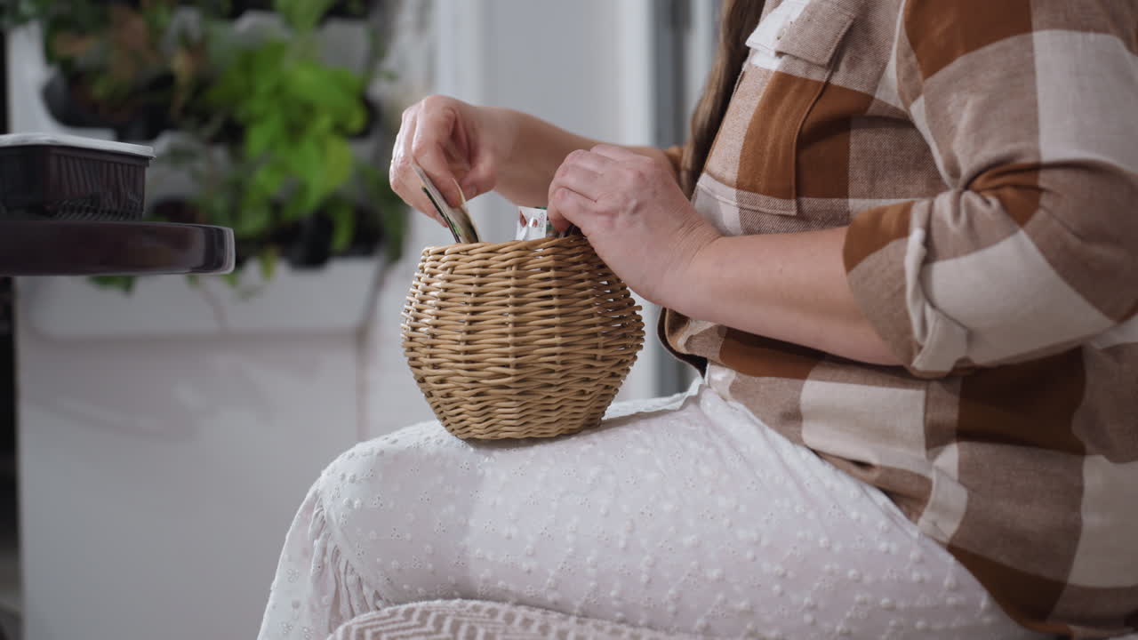 Lower view of woman seated on cozy sofa near round glass table arranging paper seed card inside small woven basket on lap surrounded by potted greenery against white brick wall backdrop