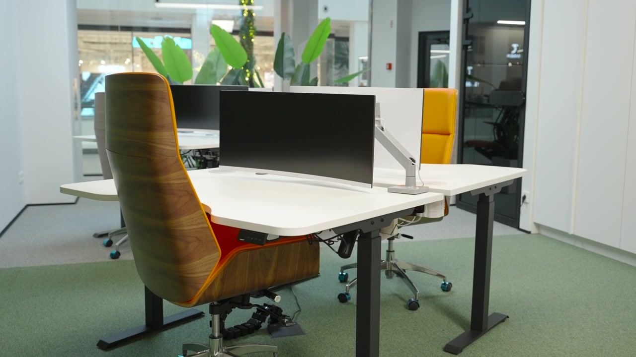 Espacio de trabajo moderno y luminoso con un monitor negro grande sobre un escritorio blanco, sillas ergonómicas, plantas de interior y luz natural que entra por amplios ventanales