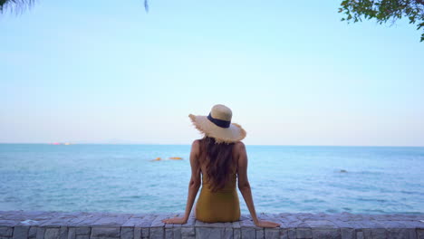 Rückansicht-Einer-Sitzenden-Frau-Mit-Langen-Haaren-Und-Einem-Strohhut,-Die-Den-Meerblick-An-Einem-Hellen-Sonnigen-Tag-Bewundert