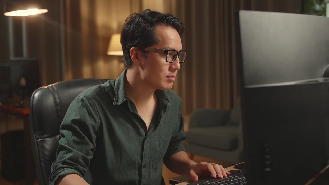 Asian Male Works His Personal Computer. He Turns And Warmly Smiles Into The Camera, He Works At Night
