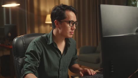 asian male works his personal computer. he turns and warmly smiles into the camera, he works at night