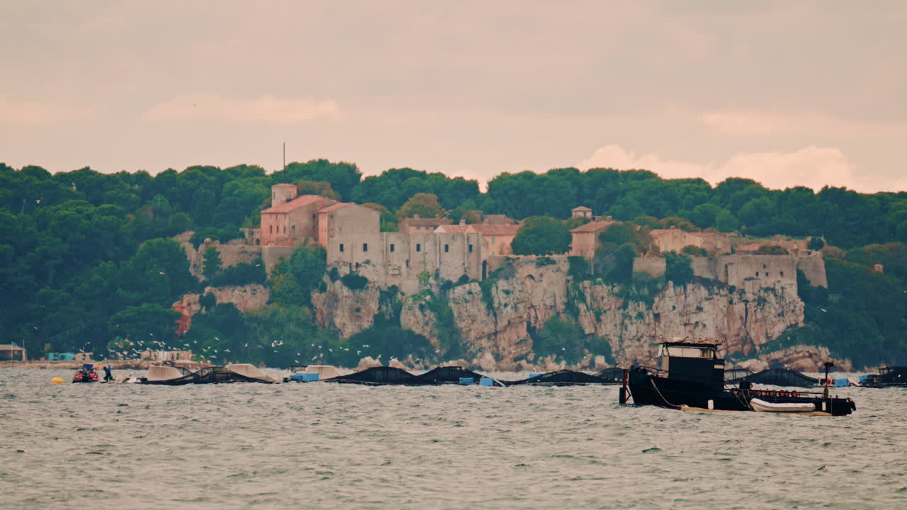 Distant view of a historic fortress on a rocky coastline surrounded by dense greenery, with boats and fish farms in the foreground