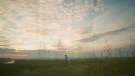 a husband and wife stand on a grassy hill beside a lake at sunset, gazing at the sky. the man, in a white shirt, hat, and jeans, holds the woman by the waist. she wears a black hat and a white dress