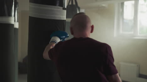 sportsman fights punching bag training in sports center. strong athlete with bald head beats hanging item in gym slow motion. skilled combat master