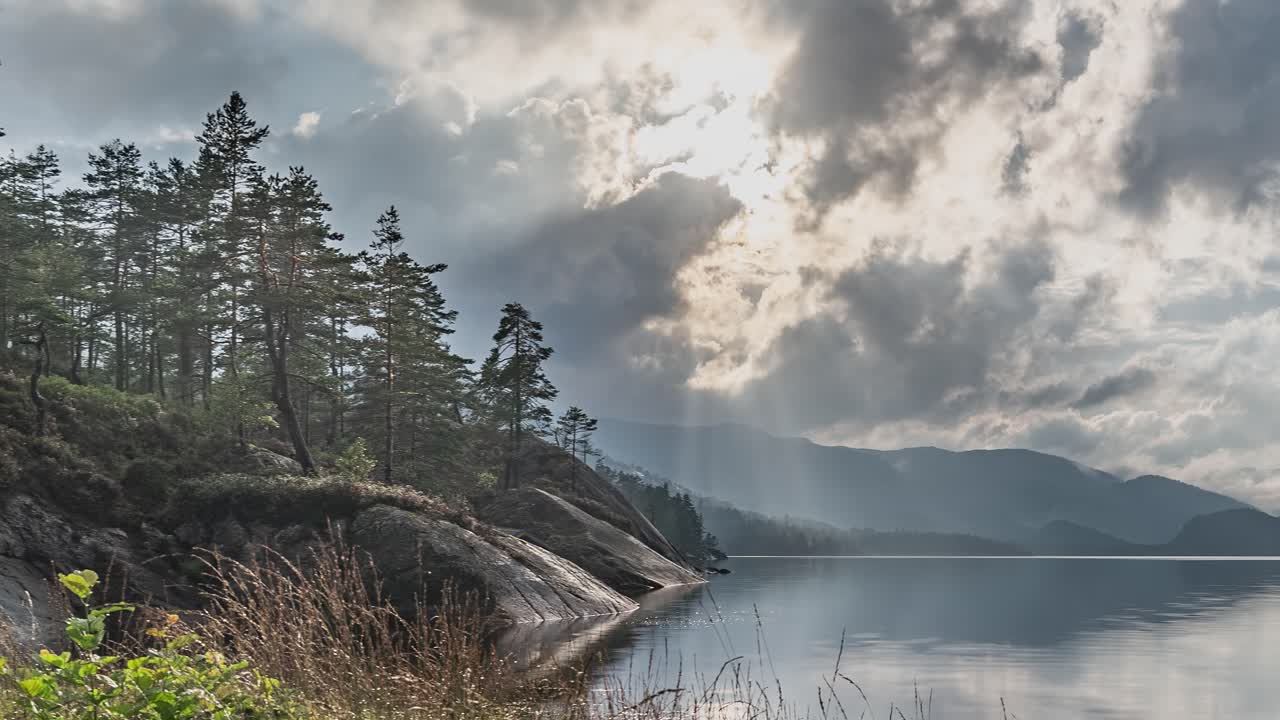 los rayos del sol atraviesan las pesadas nubes tormentosas sobre el lago tranquilo con orillas rocosas cubiertas de bosque de pinos.