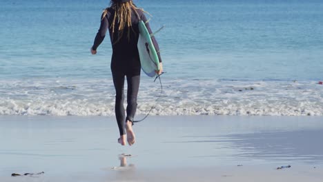 Video-Der-Rückansicht-Eines-Kaukasischen-Mannes-Mit-Dreadlocks-Im-Neoprenanzug,-Der-Ein-Surfbrett-Trägt-Und-Ins-Meer-Rennt