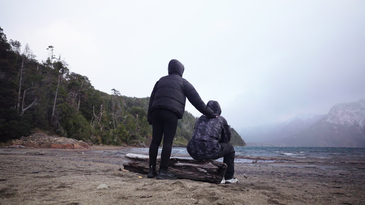 Two people in dark winter jackets share a quiet, supportive moment by the windy shore of Lake Moreno in Bariloche, Argentina, with misty skies and snowy mountains in the background