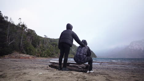 two people in dark winter jackets share a quiet, supportive moment by the windy shore of lake moreno in bariloche, argentina, with misty skies and snowy mountains in the background