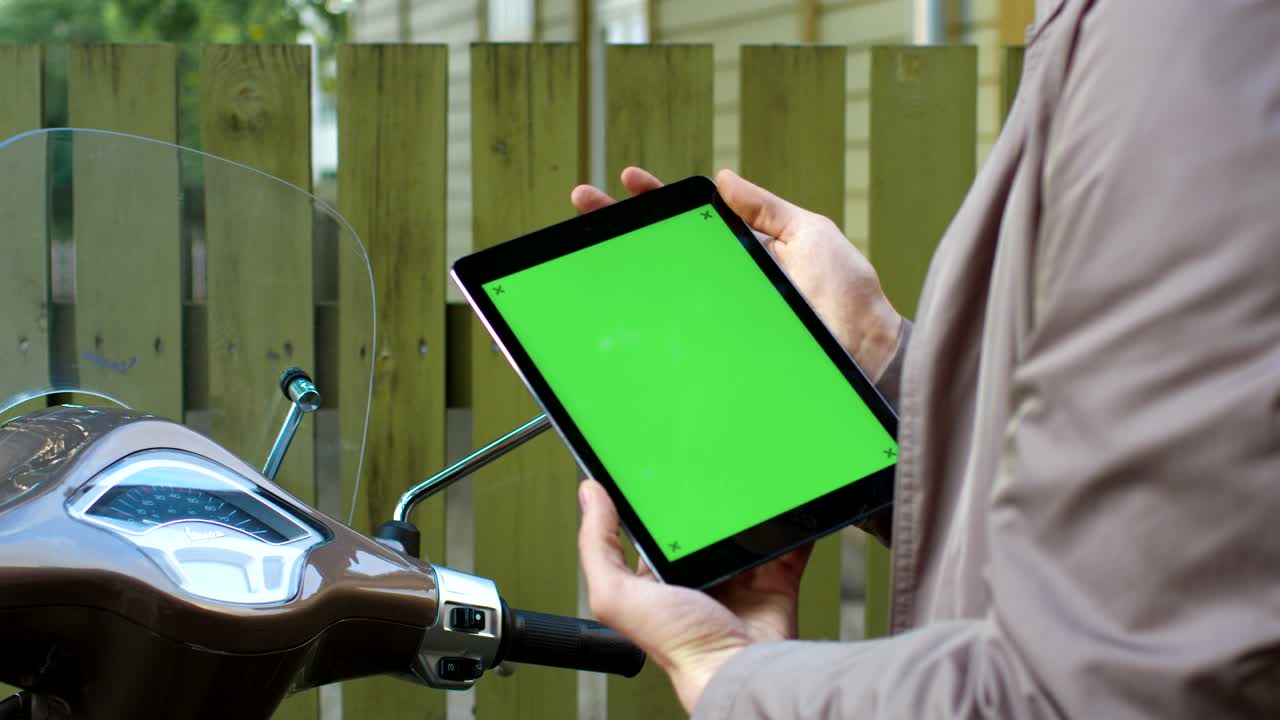un joven con un elegante abrigo beige se sienta en un scooter y sostiene una tableta con pantalla verde.