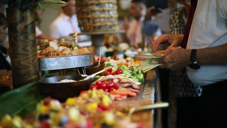 smooth shot of a line of people at a crowded event going through a food line and grabbing a variety of food including meats, cheeses, bread, fruits, and vegetables