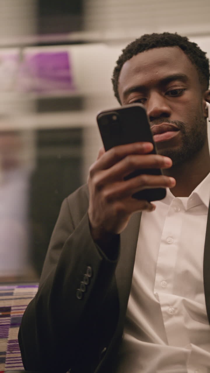 Vertical Video Of Young Businessman In Suit Commuting To Work Sitting In London Underground Tube Train Wearing Wireless Earbuds To Stream From Mobile Phone Shot In Real Time 2