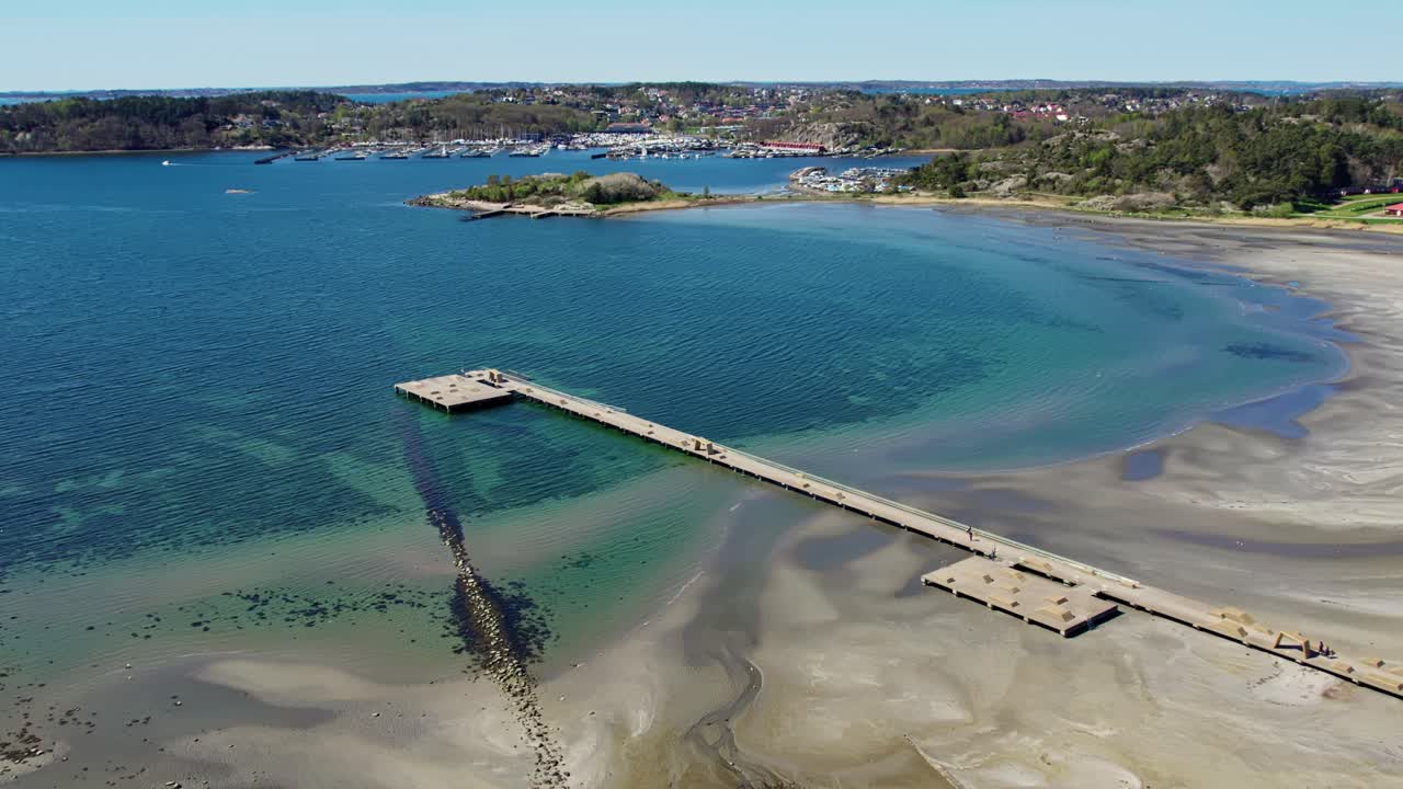 Aerial zoom over Askimsbadet beach near Gothenburg, Sweden. Features sandy shoreline, shallow waters, and a 259-meter pier reaching into the blue sea, framed by forested coast and a nearby harbor