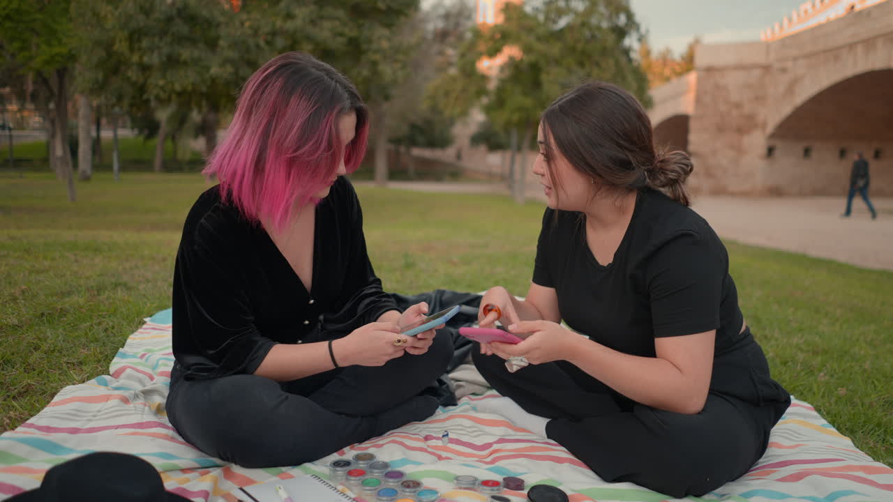 Two young women having a conversation in a park, using their smartphones while sitting on a blanket