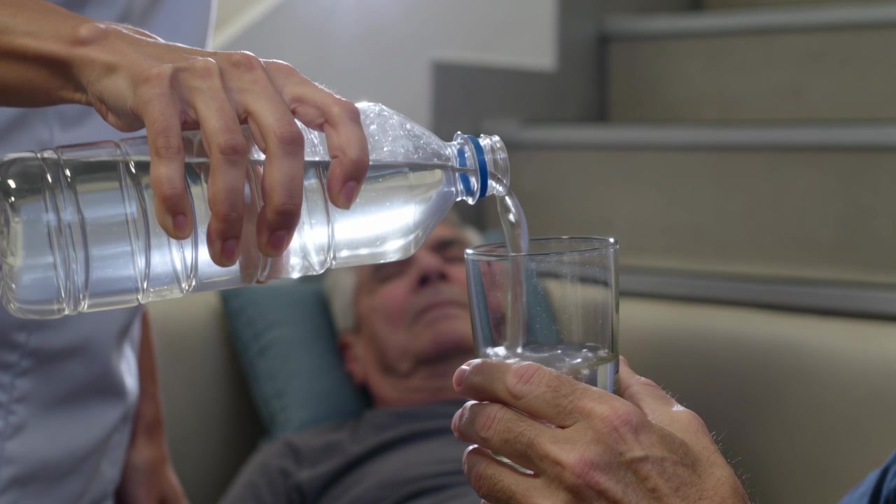 Cuidador con uniforme médico trayendo una botella, vertiendo agua en un vaso en una silla con almohada para hidratar a una persona mayor.
