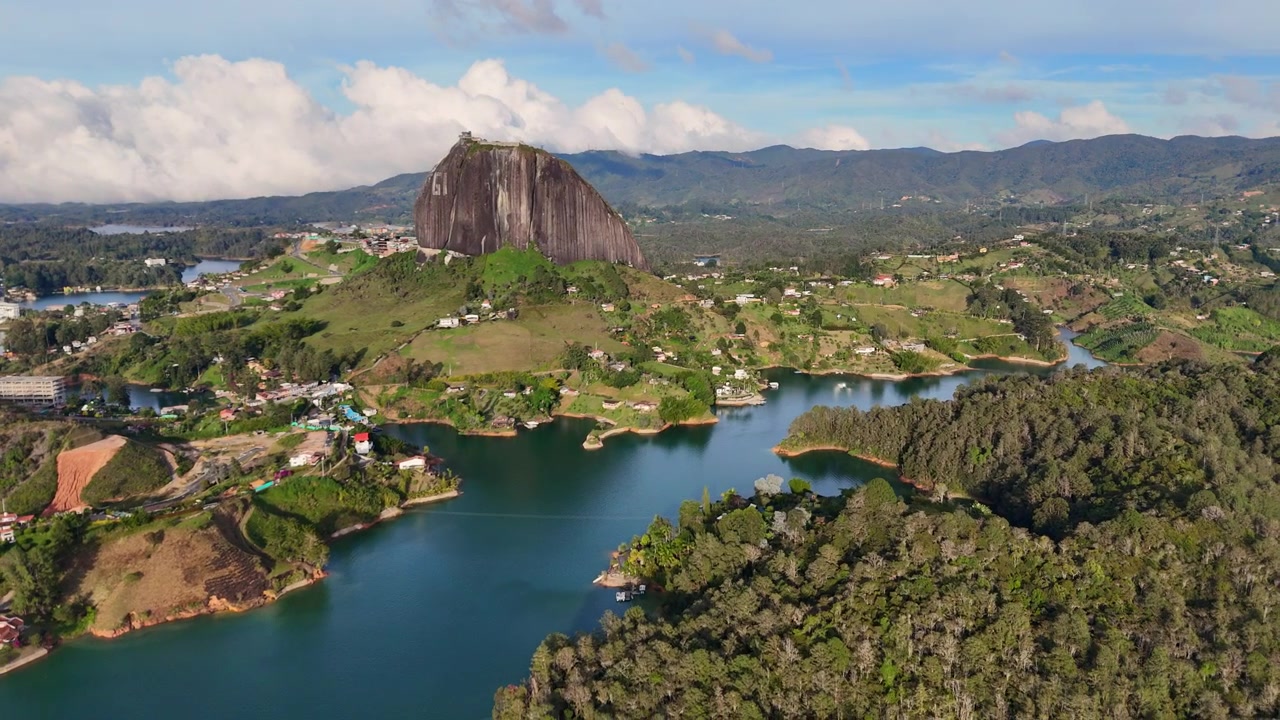Uma rotação (órbita) cinematográfica de drone, mostrando a icônica Piedra del Peñol cercada por lagos azul-turquesa e exuberantes montanhas andinas verdes em Guatapé, Antioquia, Colômbia.
