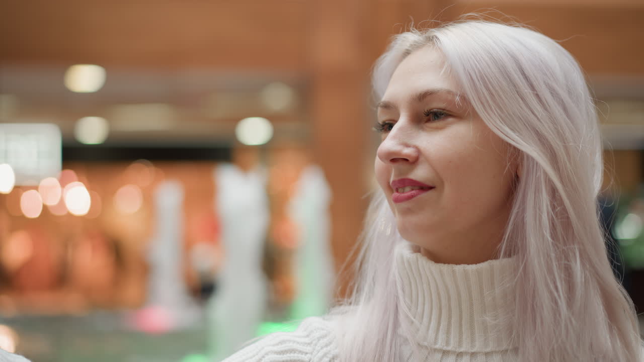 Youthful woman seated beside mall water fountain adjusts hair and smiles into mobile phone for selfie as water jets sparkle in background, modern interior ambience and joyful expression captured