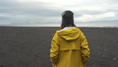 Mujer-Con-Un-Impermeable-Amarillo-Caminando-En-Una-Playa-De-Arena-Negra-En-Hvitserkur,-Vatnsnesvegur-Ubicada-En-Islandia-1