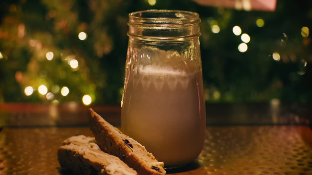 Primo piano di latte versato in un barattolo di vetro con biscotti cantucci di lato, pronto per Babbo Natale, luci dell'albero di Natale sullo sfondo