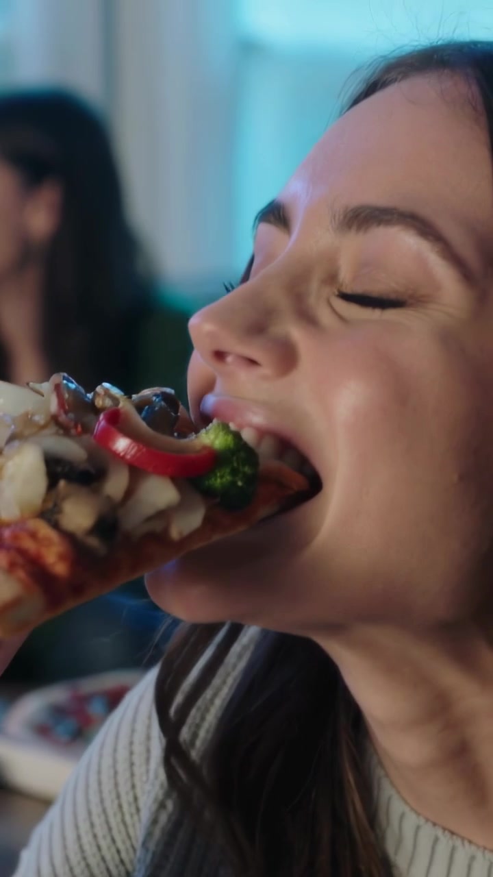Video vertical: Mujer sonriente con suéter gris llevándose un trozo de pizza a la boca, mordiendo, masticando en casa.